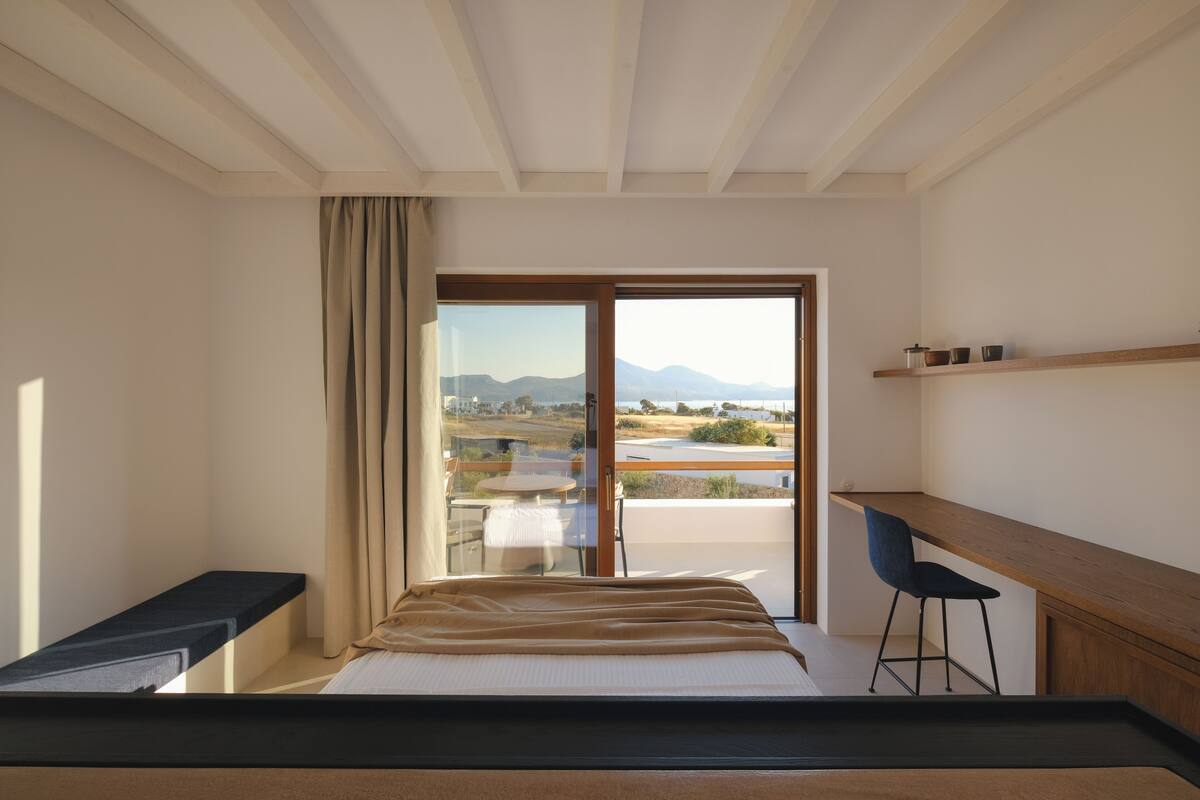 A bedroom with a bed, a desk, and a chair. There is a window with a view of mountains and a landscape.