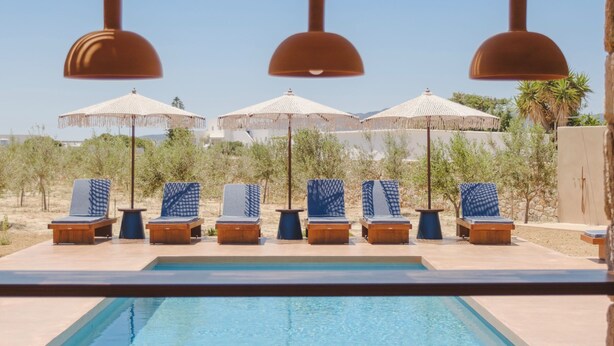 Un área junto a la piscina con sillones de descanso y sombrillas, un cielo azul despejado y una pérgola de madera.