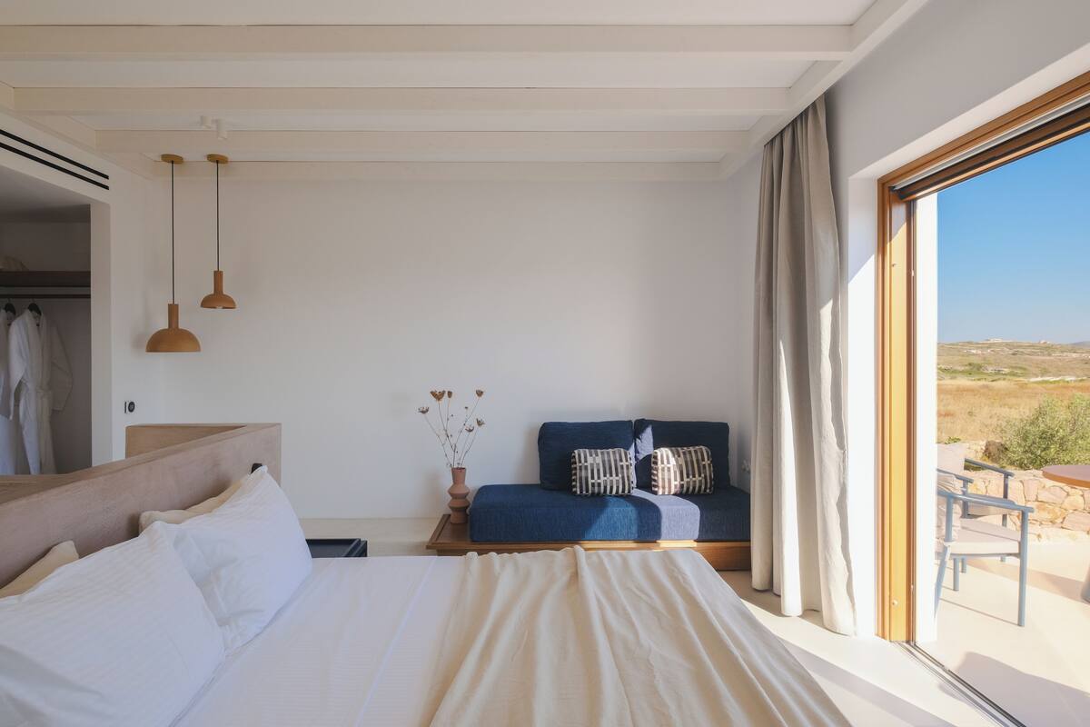 A bedroom with a bed, a sofa, a window with a view of the countryside, and a hanging light fixture.