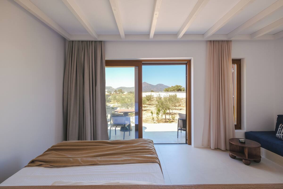 A bedroom with a bed, a small table, a sofa, and a sliding glass door leading to an outdoor area.