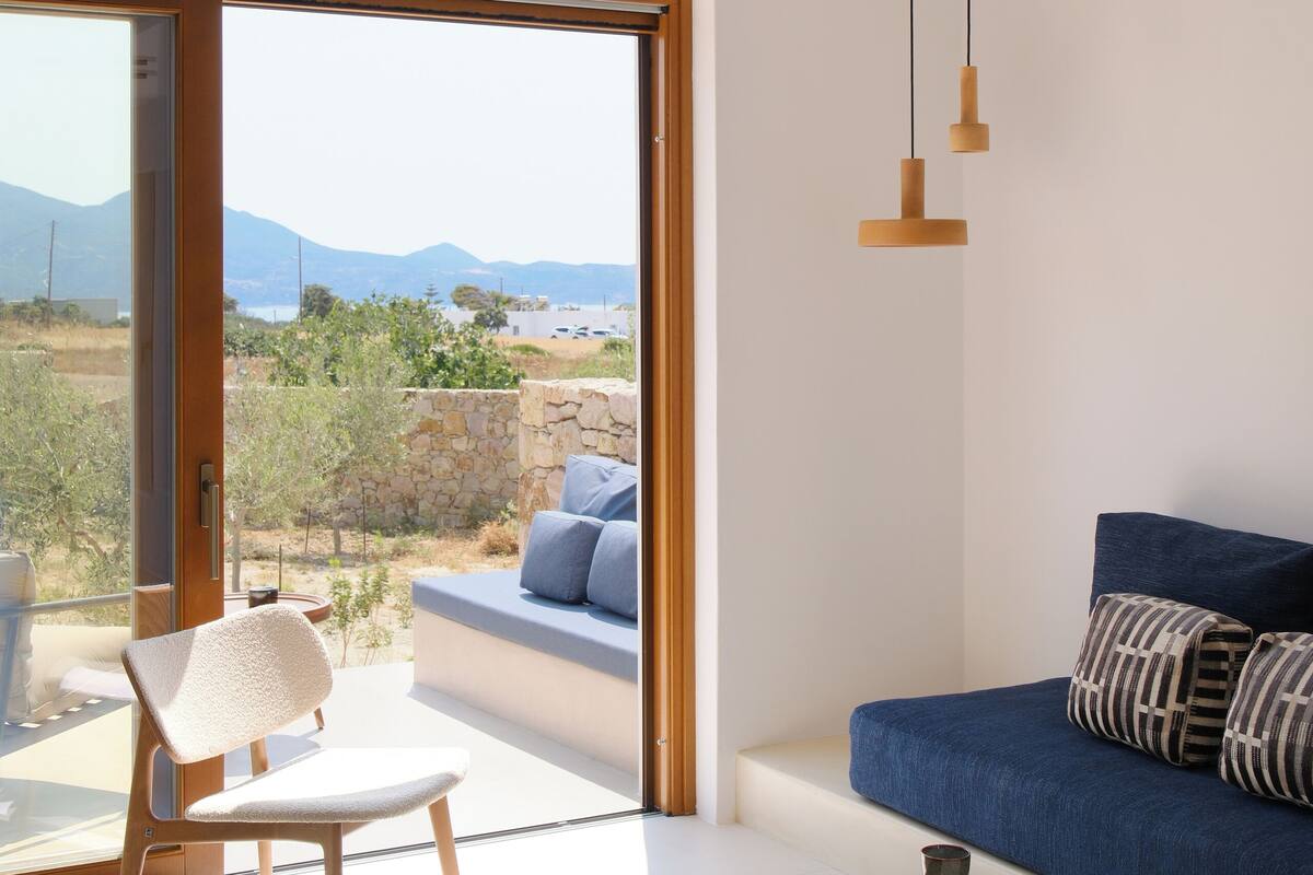 A modern living room with a large sliding glass door, a blue sofa, a wooden chair, and a round wooden side table with a book and a cup.