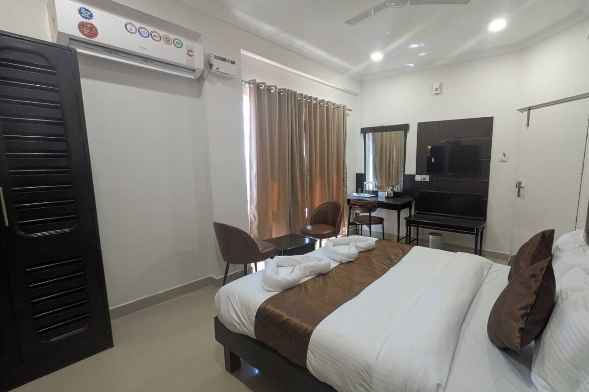 A hotel room with a bed, two chairs, a desk, a TV, and an air conditioning unit.