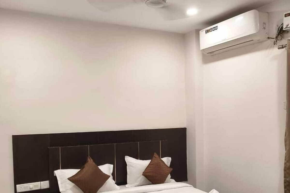 A neatly made bed with white and brown bedding, a dark headboard, a bedside table, and an air conditioning unit on the wall.