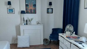 A bedroom with a bed, a dresser, a chair, a desk, a blue rug, a radiator, a painting, and a curtain.