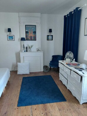 Ein Schlafzimmer mit Bett, Schminktisch, Sessel, Schreibtisch, blauem Teppich, Heizkörper, einem Gemälde und einem Vorhang.