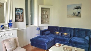A living room with a blue sectional sofa, a white coffee table, a chandelier, a fireplace, and a painting of beach chairs.