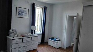 Un dormitorio con cómoda, ventilador, lámpara, ventana con cortinas, una pequeña cesta de mimbre y una puerta.