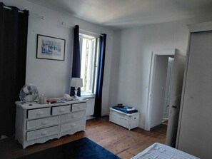 Ein Schlafzimmer mit einem Schminktisch, einem Ventilator, einer Lampe, einem Fenster mit Vorhängen, einem kleinen Korbwesen und einer Tür.