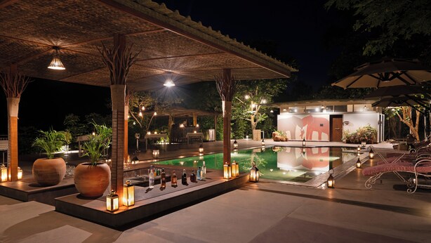 An outdoor pool area with a covered seating area, ambient lighting, and potted plants. - Brij Sone Bagh, Bandhavgarh | A Luxury Jungle Retreat (Umaria)
