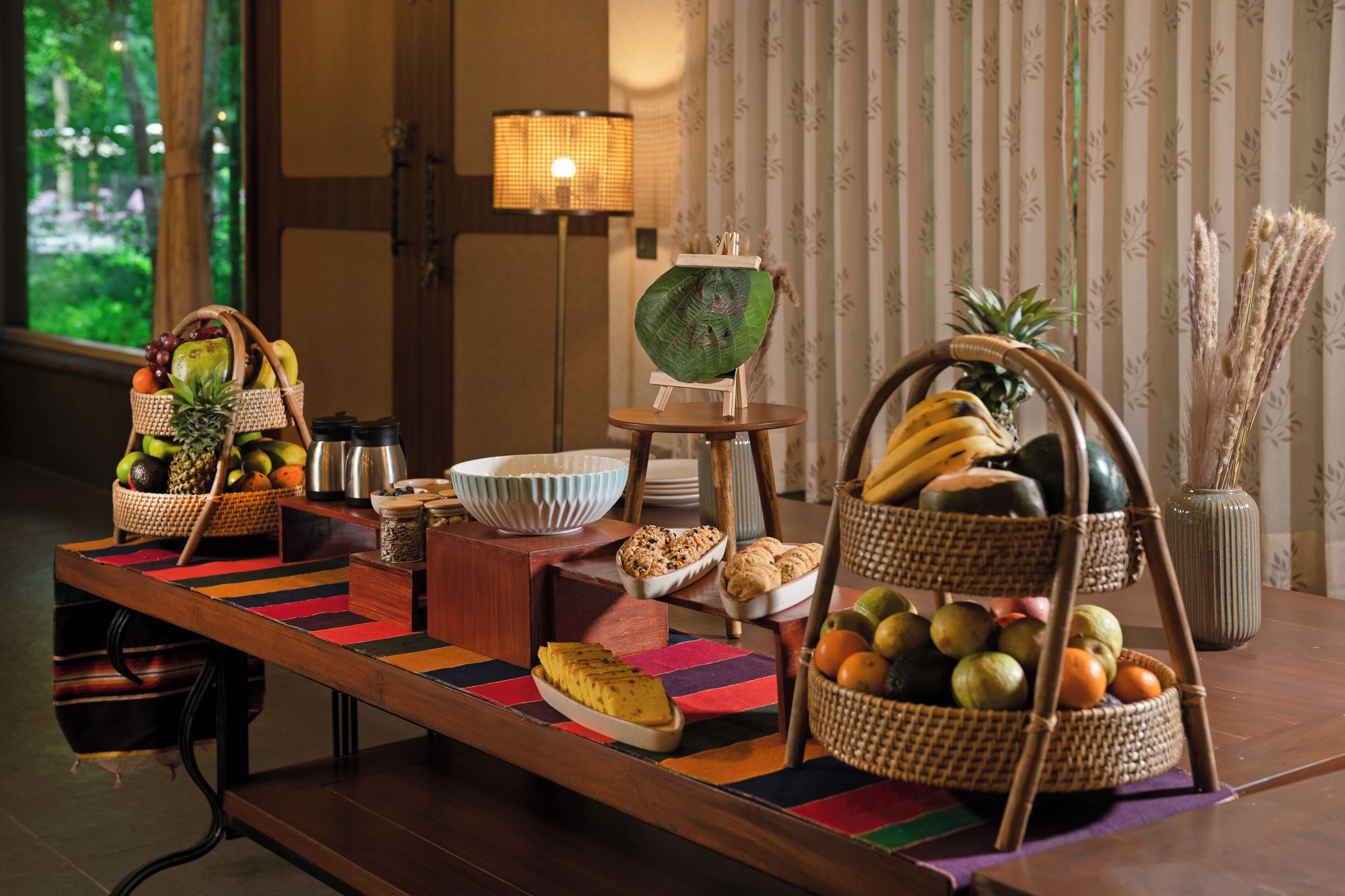 Una mesa de bufé con diversas frutas, una lámpara y un florero con plantas secas.