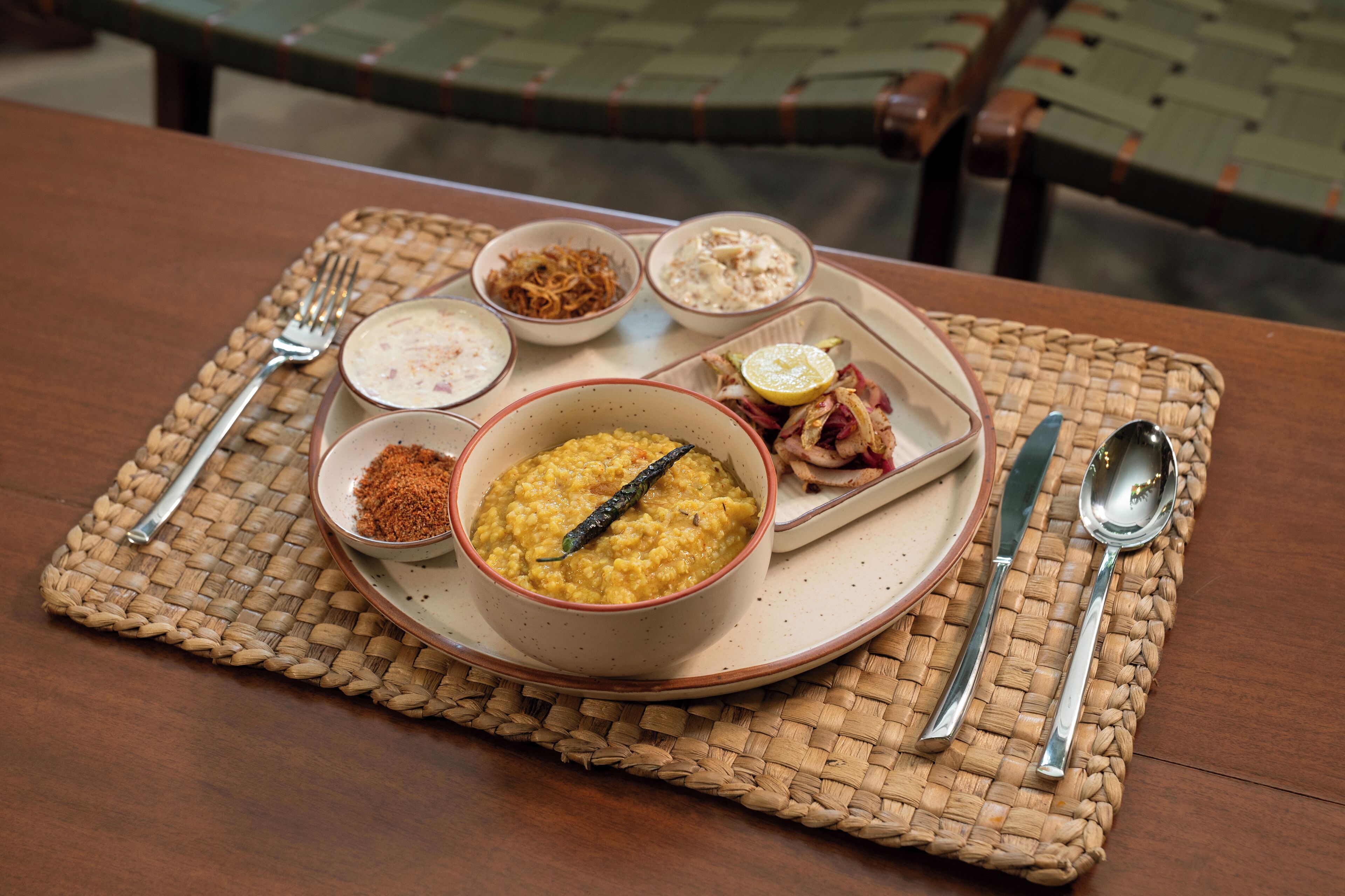 A table with a woven placemat, a plate of food including a bowl of dal, a bowl of chutney, and a small dish of pickled vegetables.
