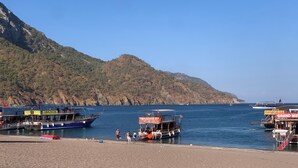 Une zone côtière où des bateaux sont amarrés à un quai, avec une chaîne de montagnes en arrière-plan et un ciel dégagé.