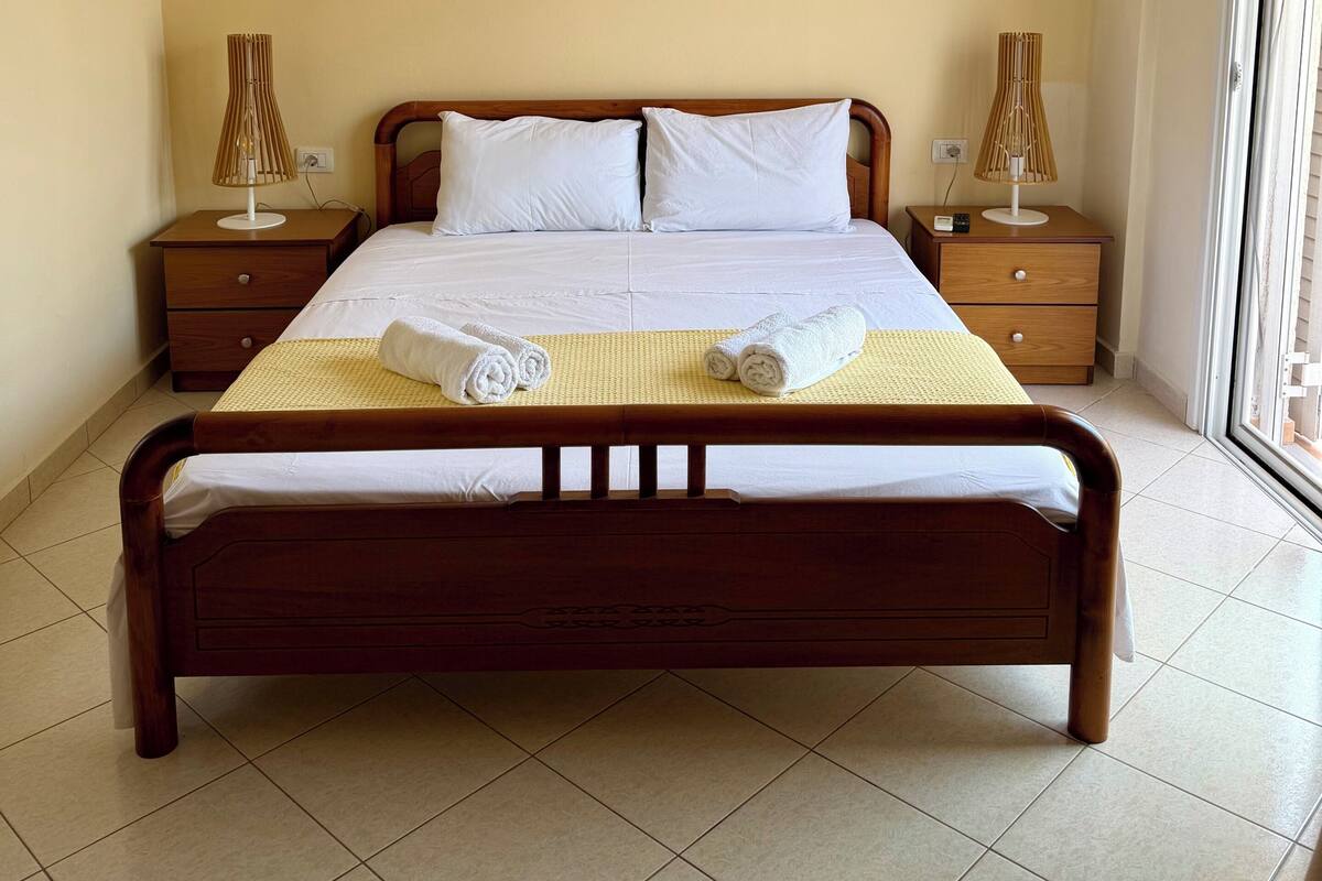A neatly made bed with white linens, two bedside tables with lamps, and an air conditioning unit on the wall.