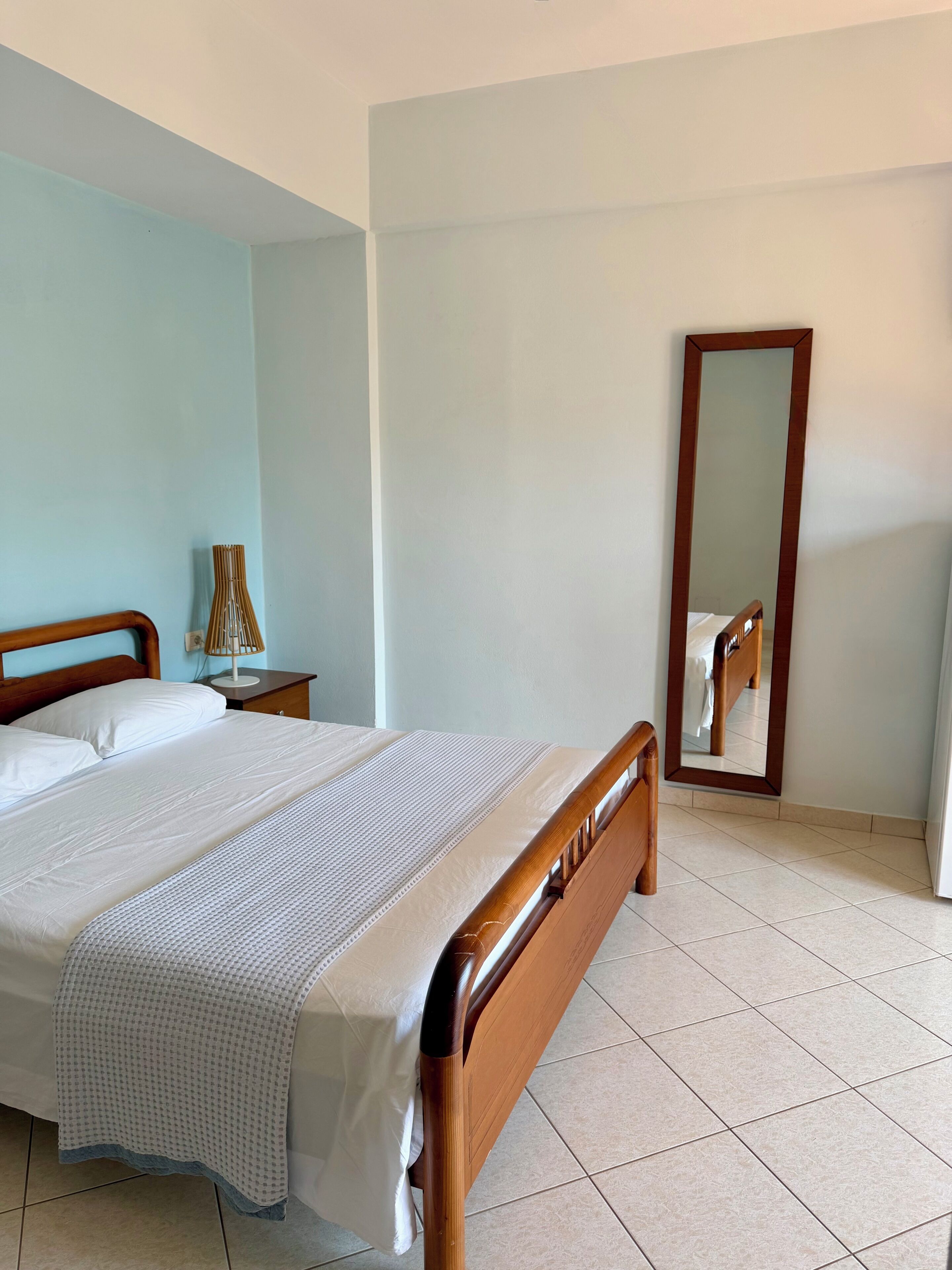 A neatly made bed with a wooden headboard, a bedside table with a lamp, and a full-length mirror on the wall.