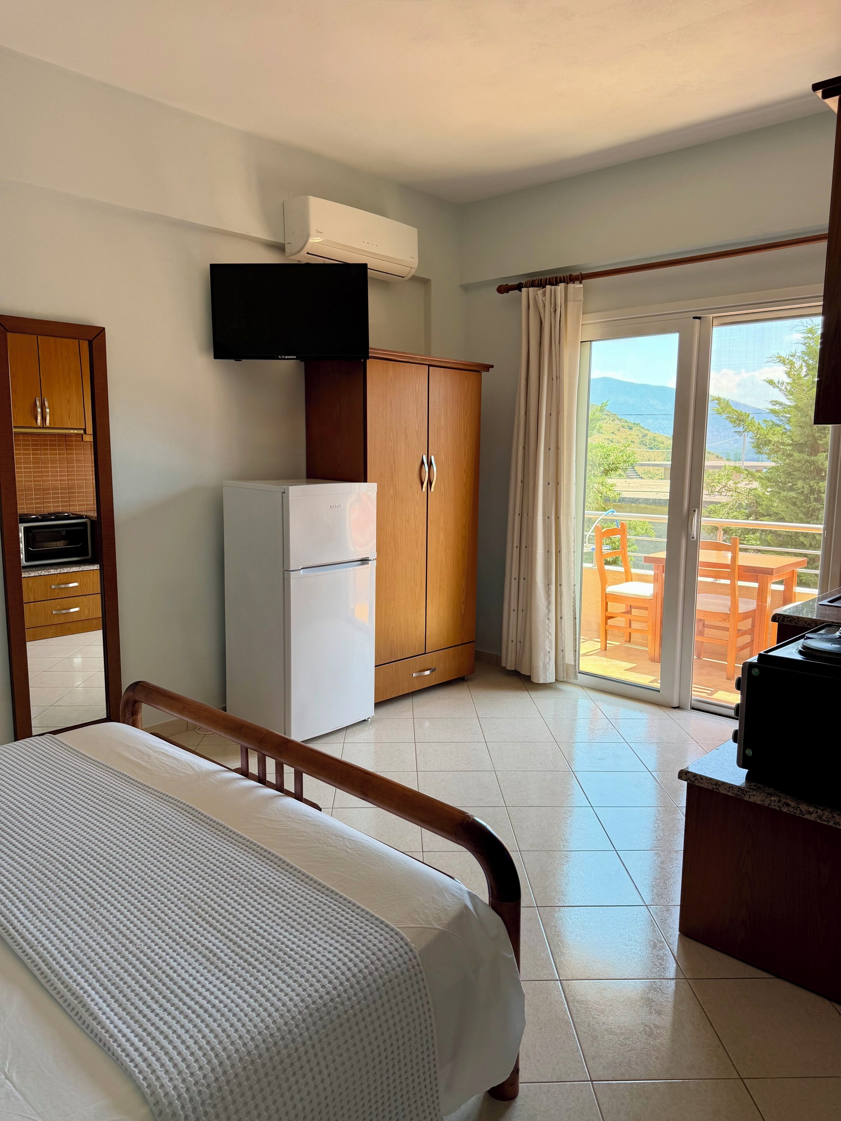 A bedroom with a bed, a wardrobe, a refrigerator, a television, and a balcony with a table and chairs.