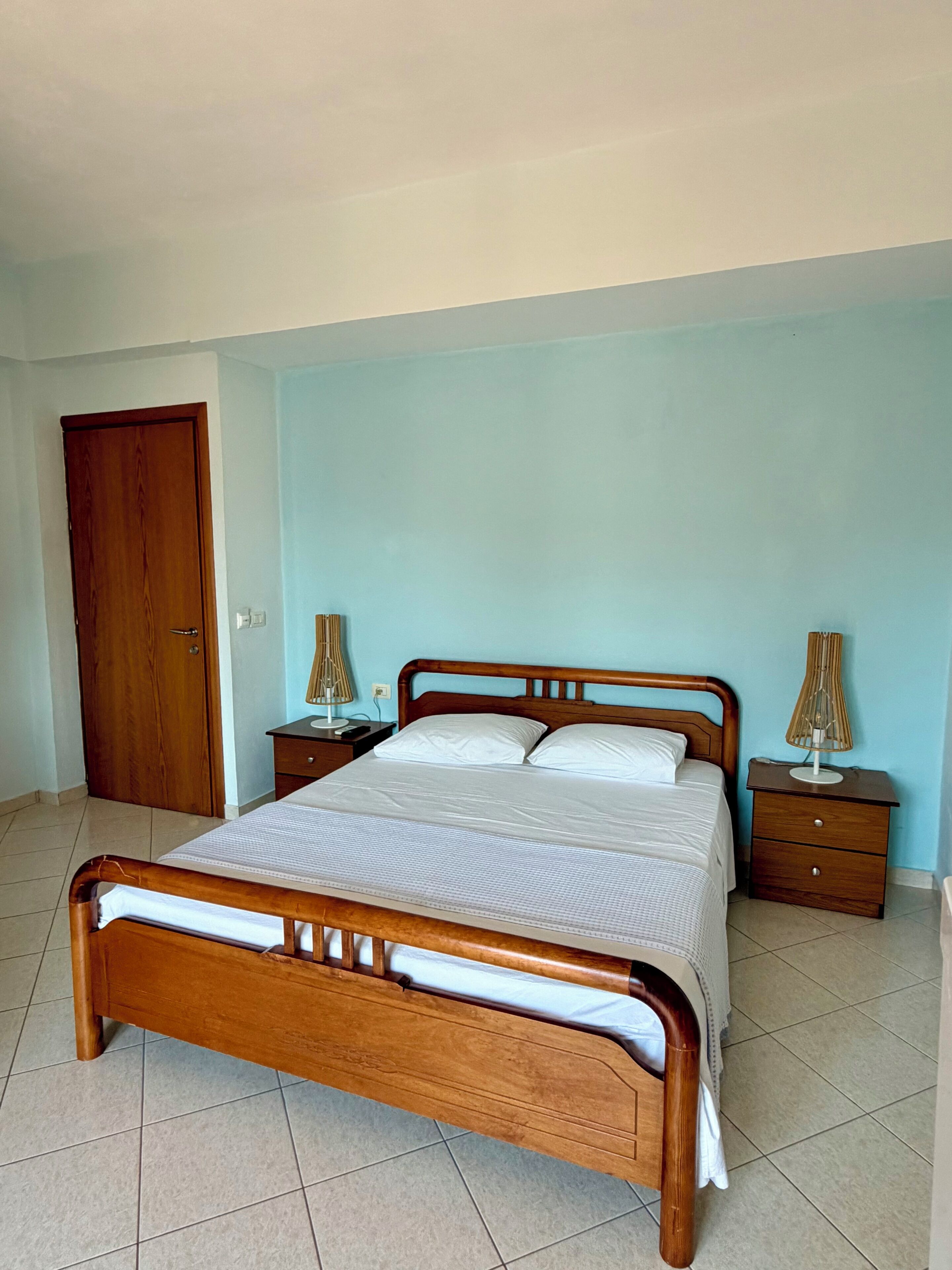Une chambre à coucher avec un lit en bois, deux tables de chevet, une commode, une porte en bois et un mur bleu clair.