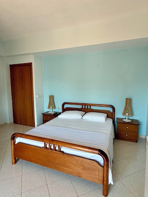 A bedroom with a wooden bed, two bedside tables, a nightstand, a wooden door, and a light blue wall. - Mihasi Apartments (Ksamil)