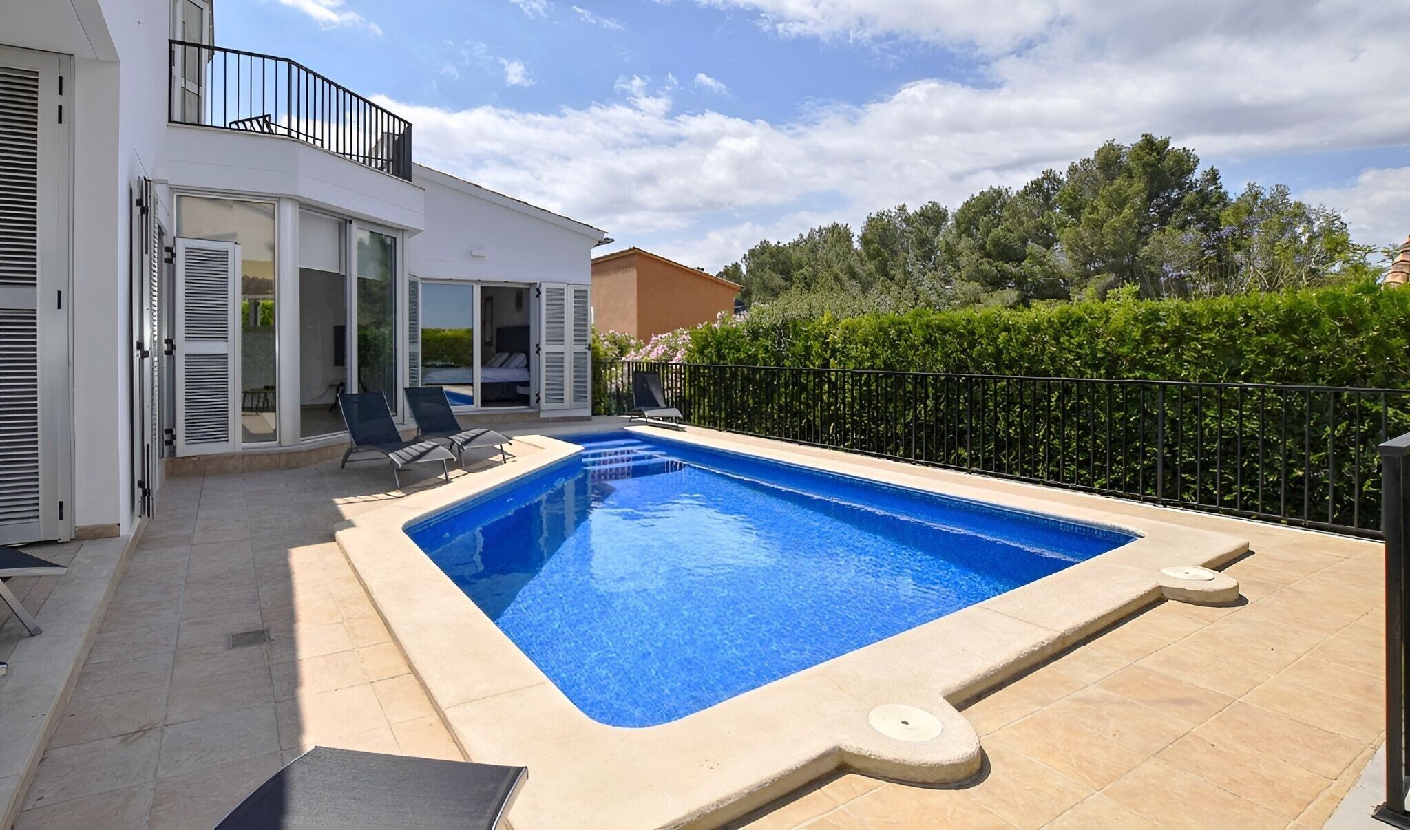 A rectangular swimming pool with a tiled deck, two lounge chairs, and a glass door leading to a room.