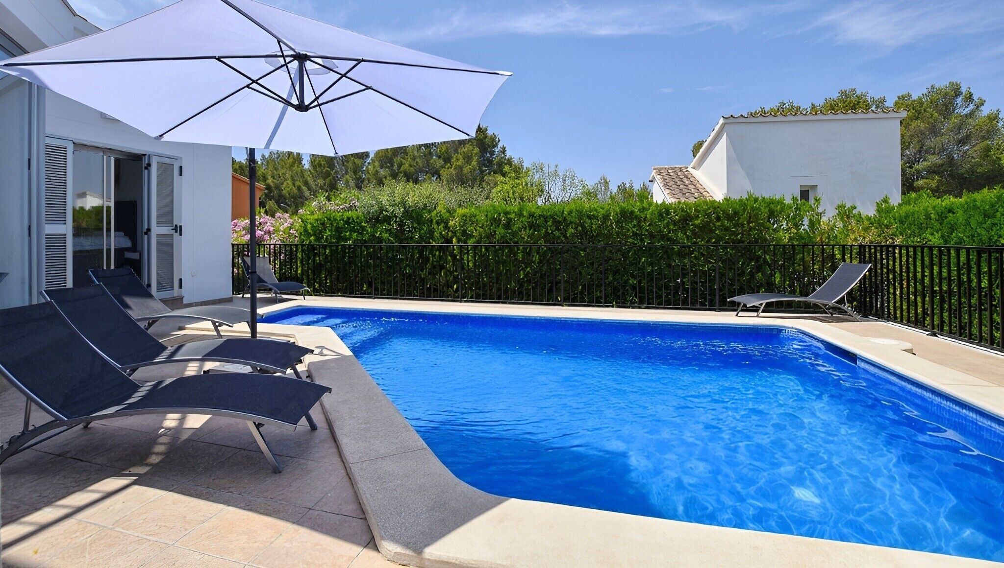 Une piscine entourée d’une clôture et de verdure, avec des chaises longues et un parasol.