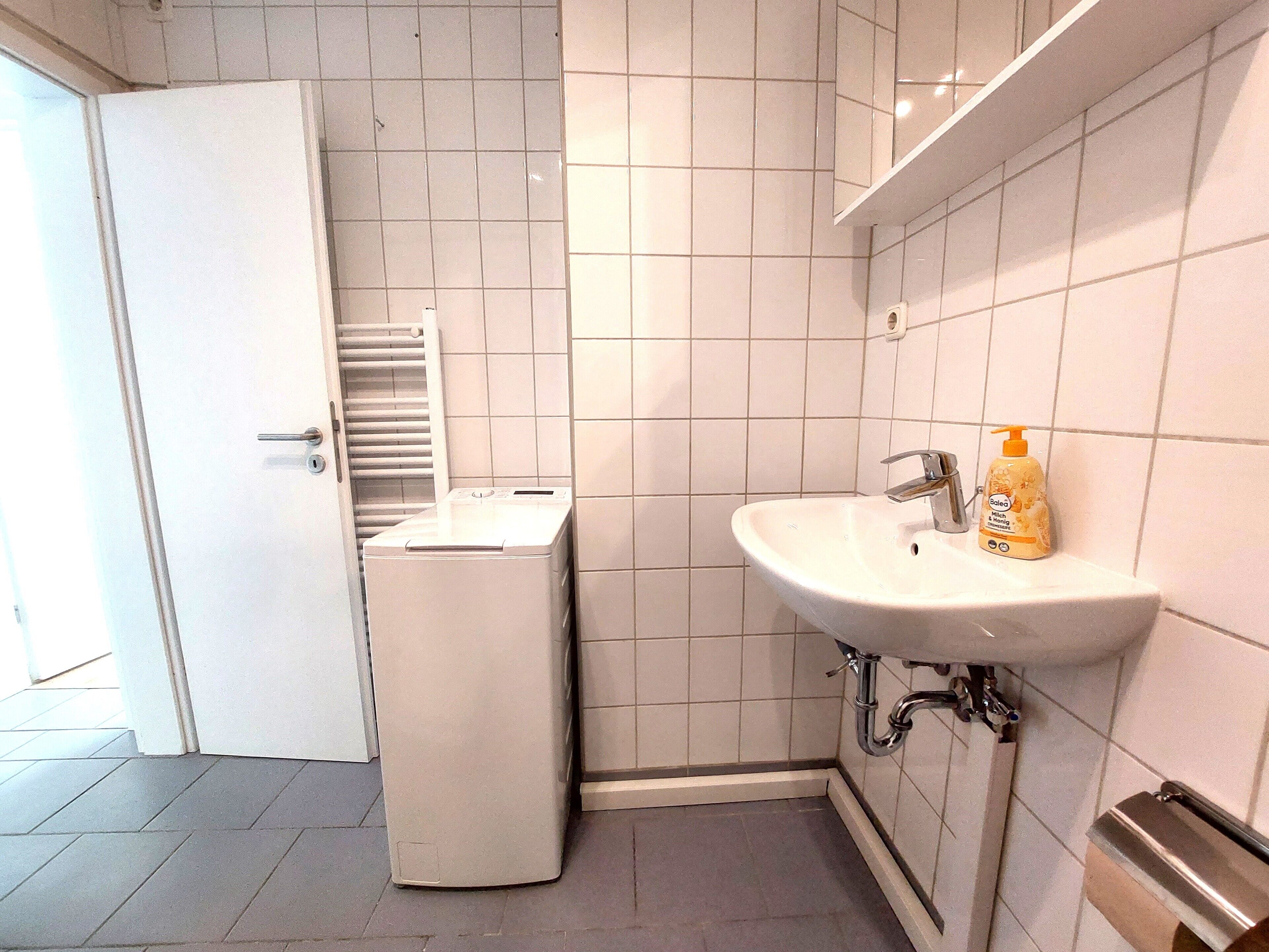 Un baño con pared de azulejos blancos, un lavamanos con dispensador de jabón y una lavadora blanca.