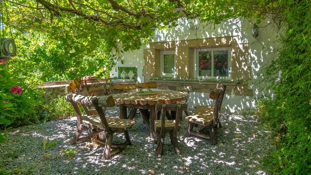 An outdoor patio with a wooden table and chairs, surrounded by greenery and a stone wall.