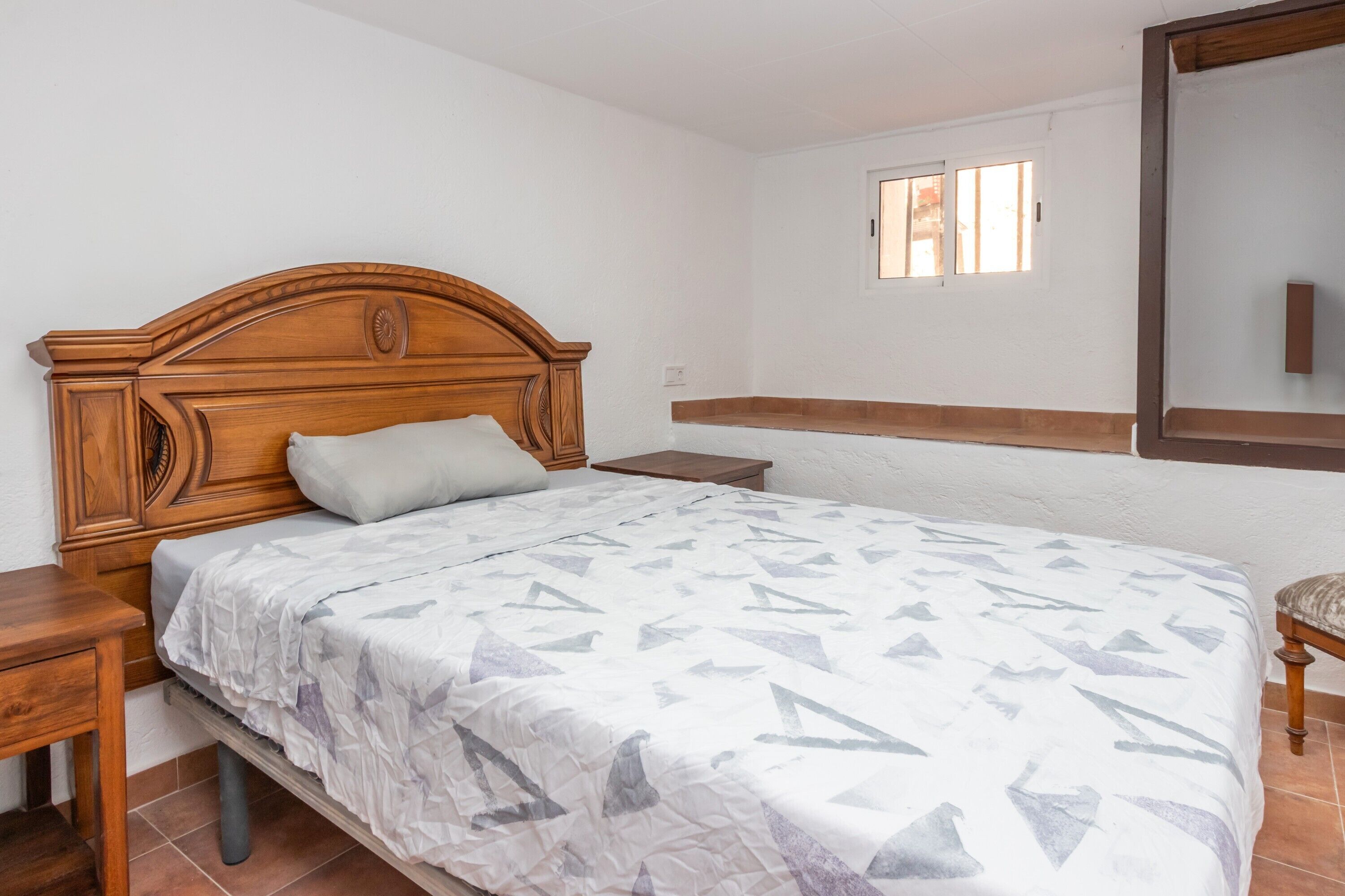 Een slaapkamer met een houten hoofdbord, een bed met een bedovertrek met een patroon, een nachtkastje, een houten stoel en een raam met jaloezieën.