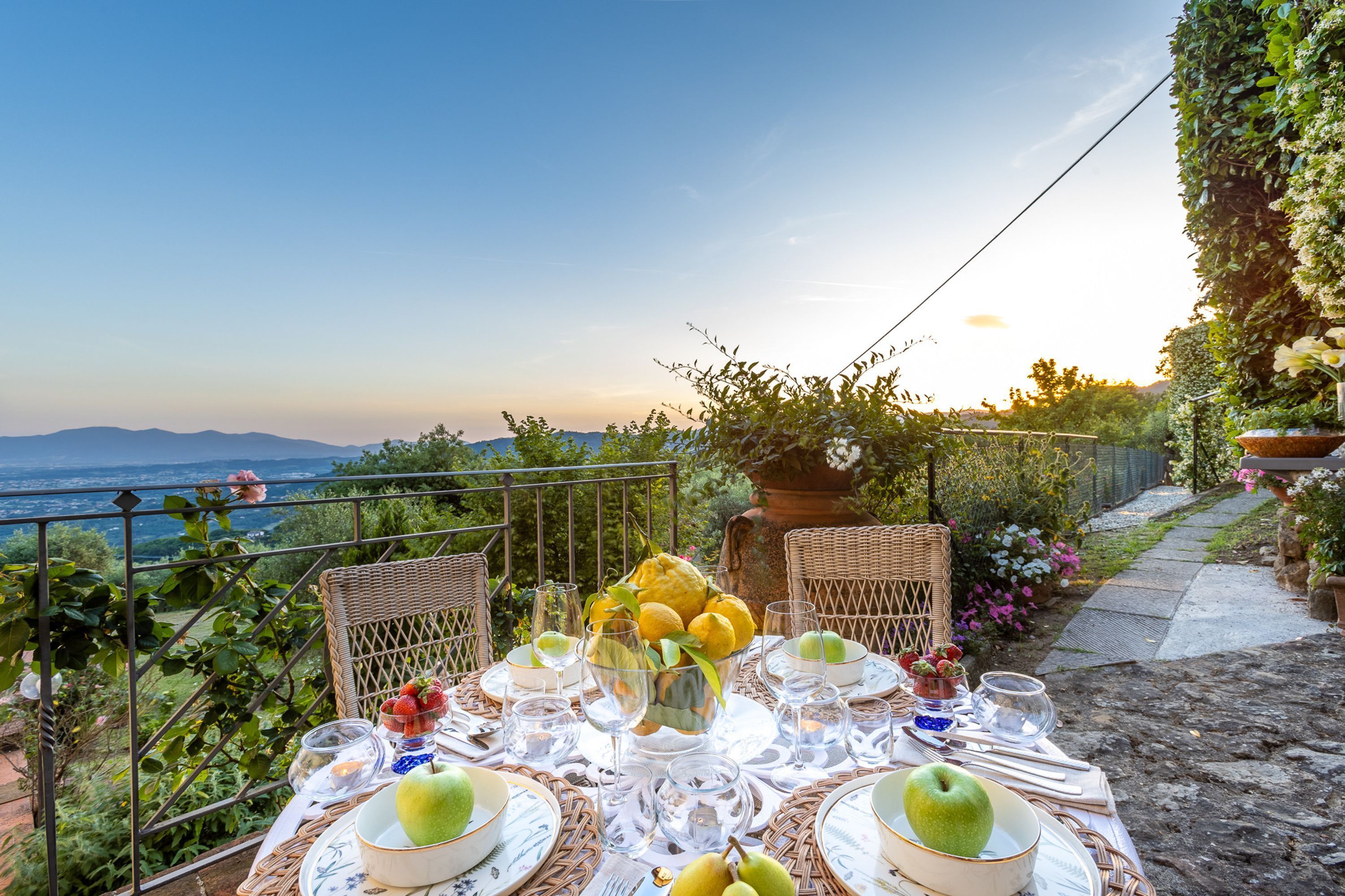 An outdoor dining table set with plates, glasses, and fruits, overlooking a scenic landscape with mountains and a body of water.