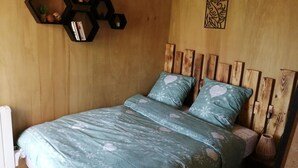 Een bed met een bloemenbedekking en kussens met een hartpatroon, een houten hoofdbord, een zeshoekig schap met decoratieve items en een kunstwerk aan de muur.