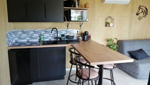 Een compacte keuken met een zwarte koelkast, een spoelbak en een houten aanrecht. Op het aanrecht staan een zwarte koffiezetapparaat en een waterkoker. Op de achtergrond staat een grijze bank met een blauw kussen.