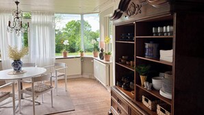 Una habitación con una mesa y sillas blancas, un mueble de madera con vajilla, y vistas a espacios verdes.
