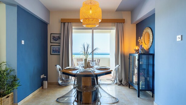 A dining area with a table set for four, clear chairs, a hanging light fixture, a view of the sea, and a cabinet with decorative items. - Bayview 2-BR Apartment in Larnaca (Larnaca)