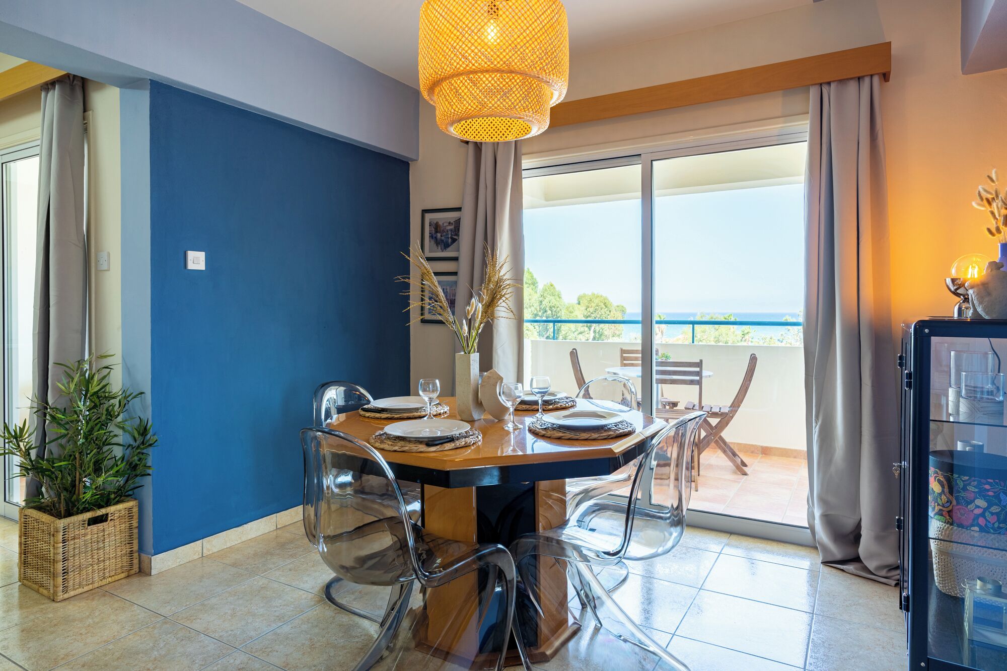 A dining area with a table set for four, clear chairs, a hanging light fixture, and a view of the outdoors through large windows.