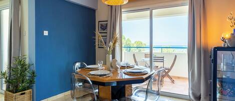 A dining area with a table set for four, clear chairs, a hanging light fixture, and a view of the outdoors through large windows.