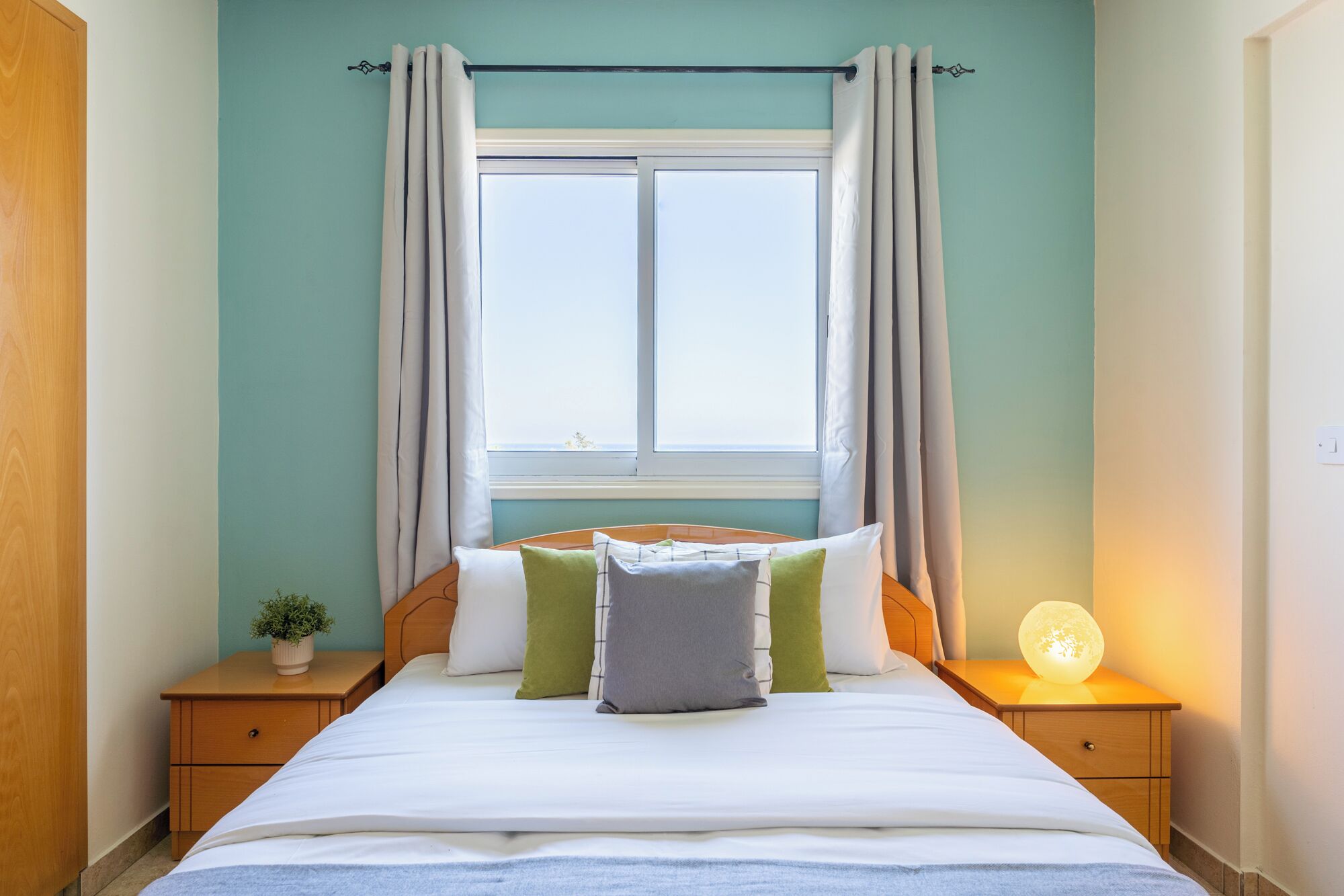 A bedroom with a bed, nightstands, a window with curtains, a bedside lamp, and a small plant.