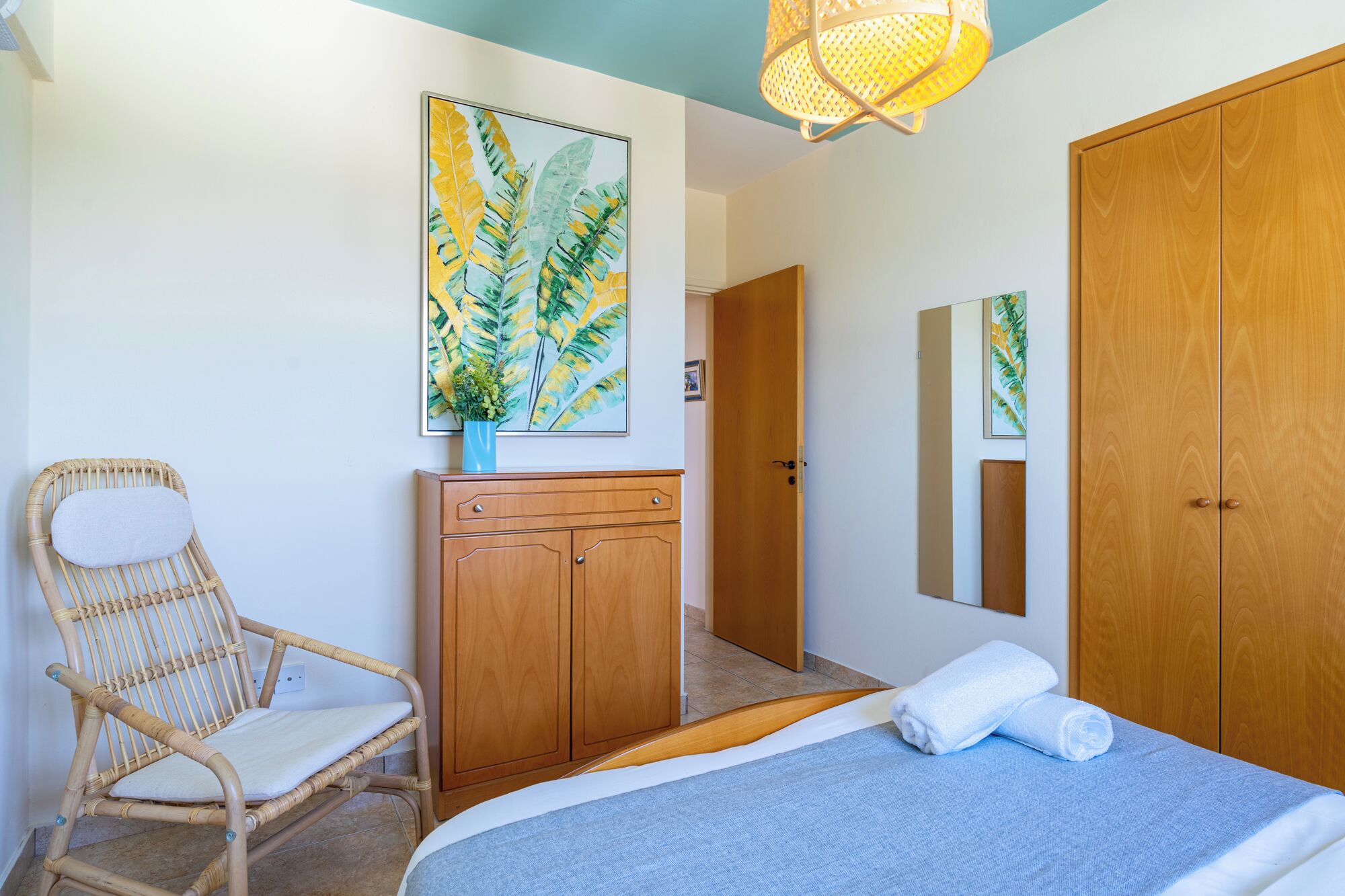 Une chambre à coucher avec un lit, une armoire en bois, une chaise en osier, un tableau et un miroir.