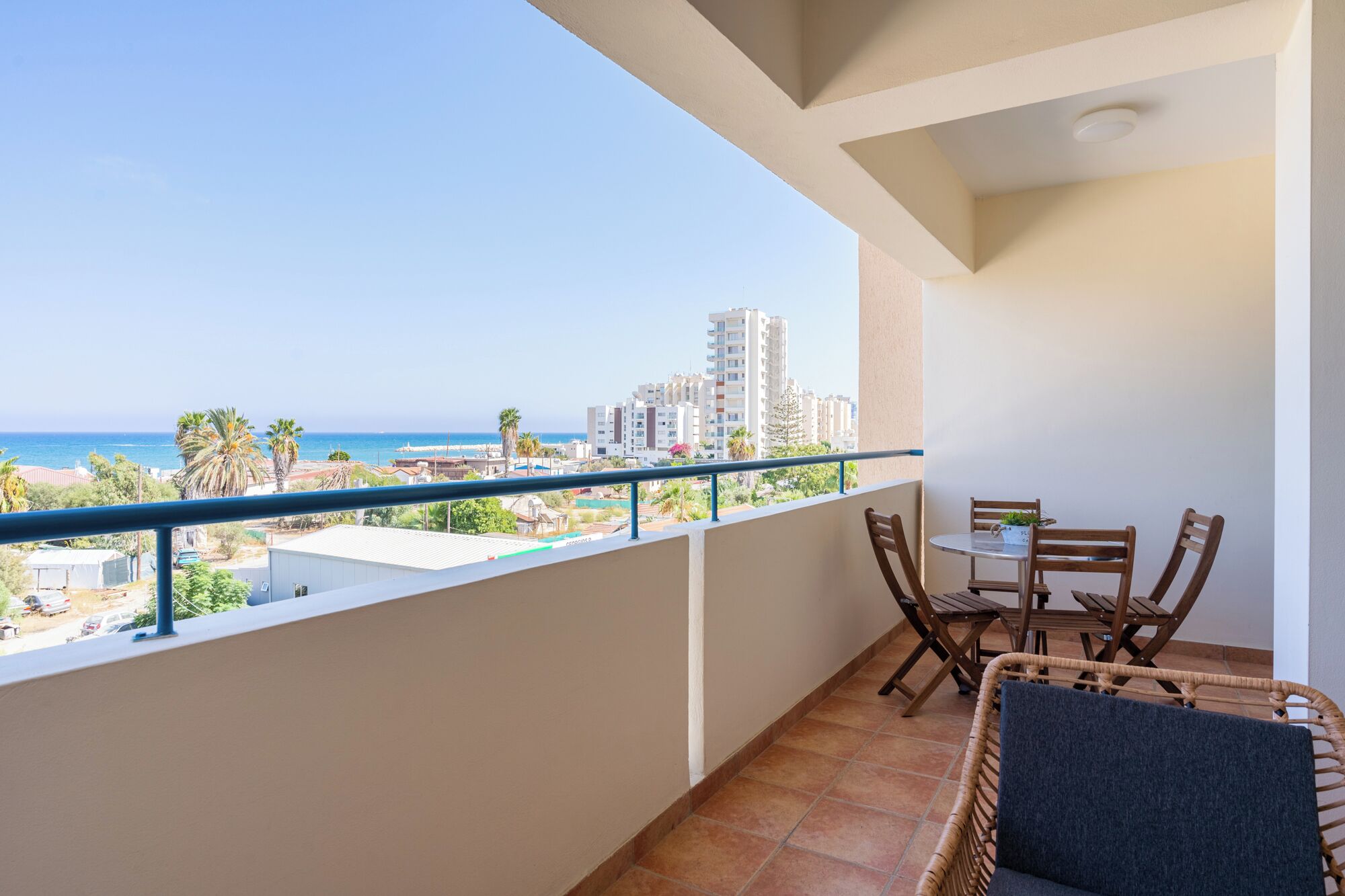 Un balcón con vista al mar y edificios, una mesa con sillas y una silla de mimbre.