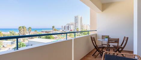 A balcony with a view of the sea and buildings, a table with chairs, and a wicker chair.