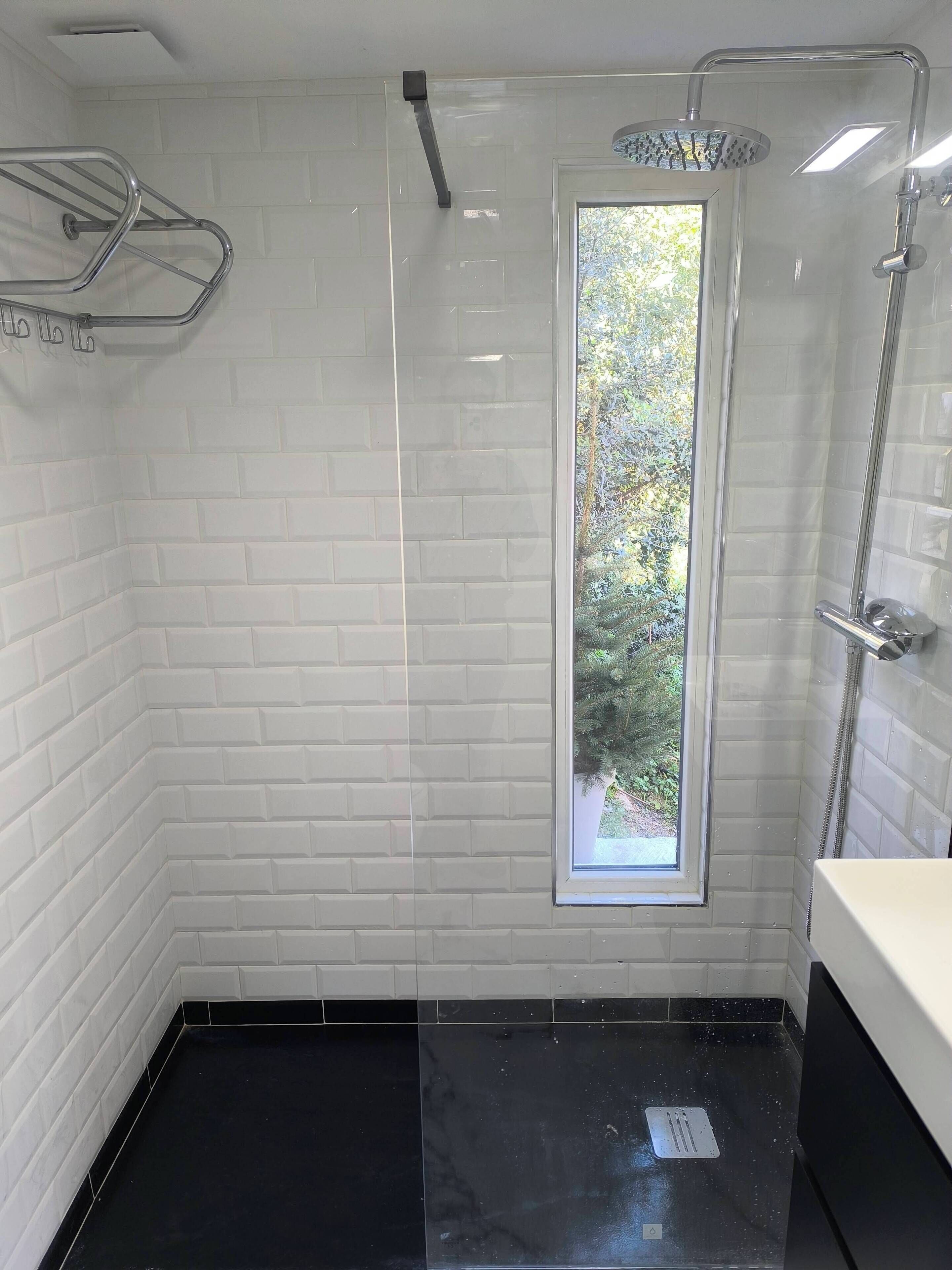 A modern bathroom with white subway tiles, a black floor, a glass shower enclosure, and a window with a view of greenery.