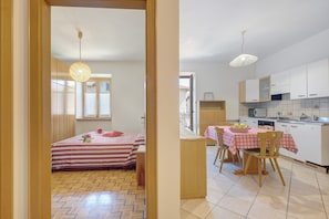 A bedroom with a bed, a bedside table, and a window. The room has wooden flooring and a hanging light fixture.