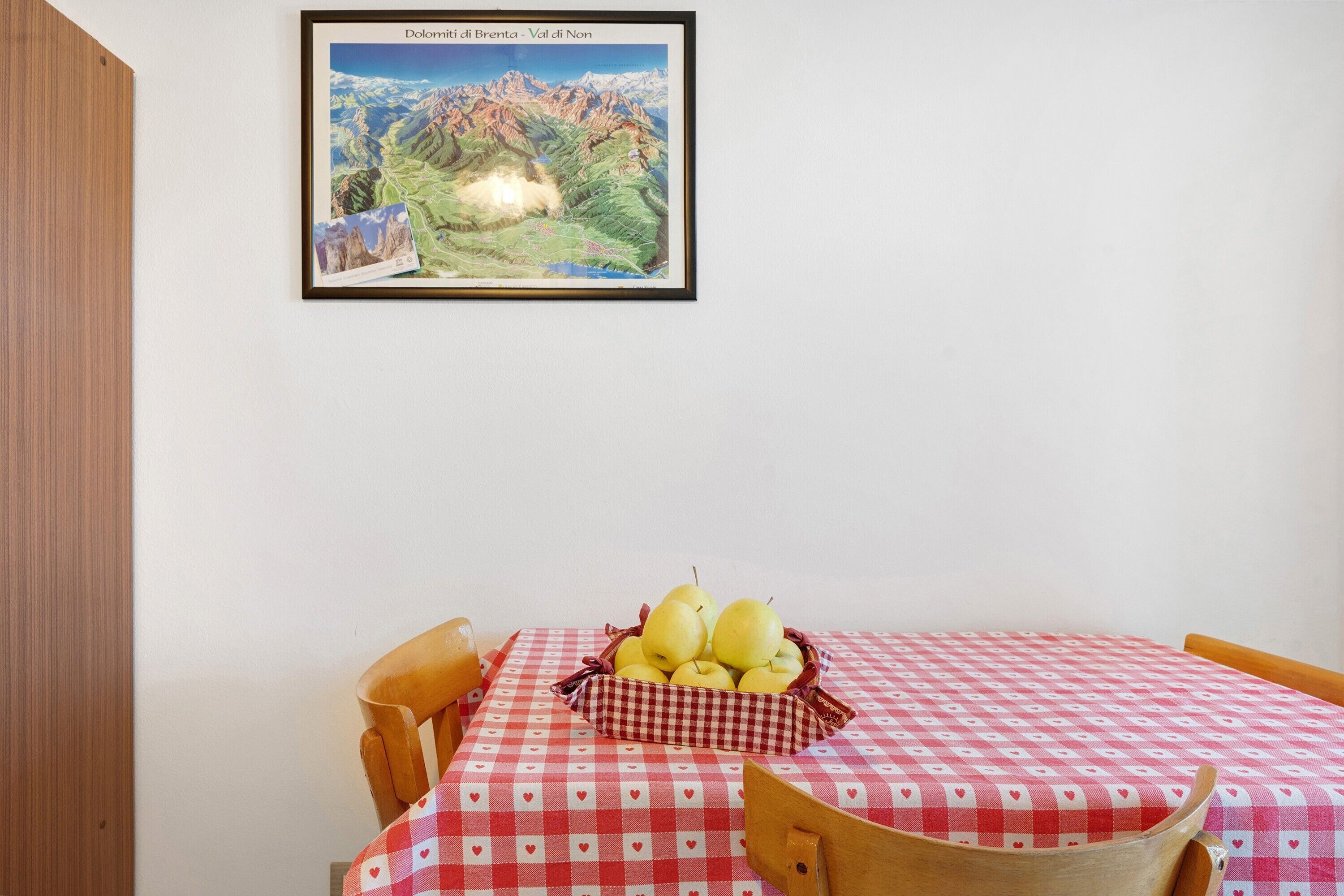 Ein Essbereich mit einem Tisch, bedeckt mit einer rot-weißen Karotisch, einer Birnenkorb und einem gerahmten Bild der Dolomiten an der Wand.