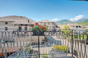 A balcony with metal railings, a table, and chairs, overlooking a residential area with houses and mountains in the background.