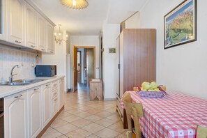 A kitchen with white cabinets, a microwave, and a sink. There is a dining area with a checkered tablecloth and a chair.