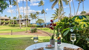 A dining table with two plates, a bottle, and two wine glasses, set against a backdrop of palm trees and a view of the ocean. - Unwind in Napili, as sunsets await (Napili)