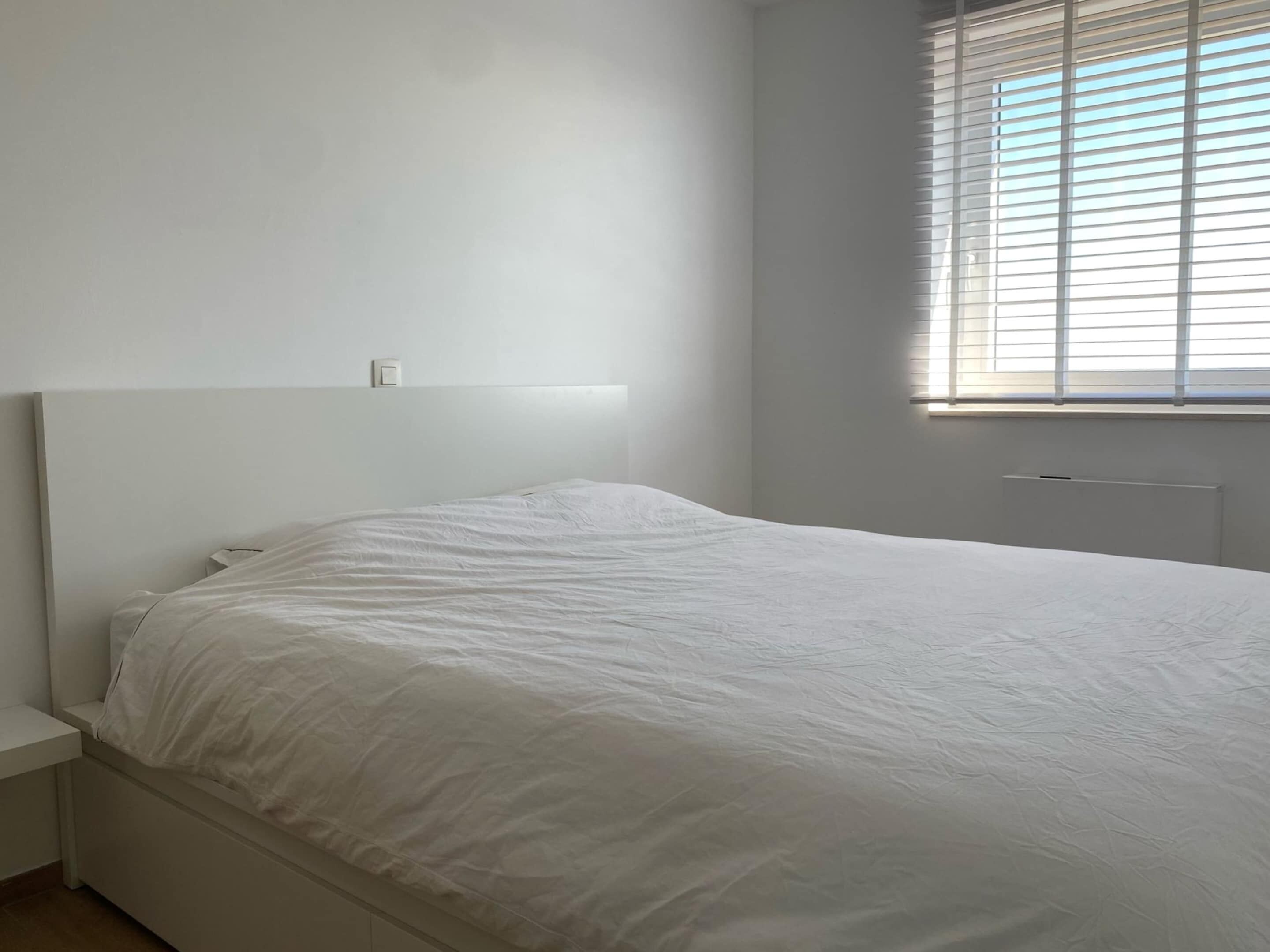 A simple, unadorned bedroom with a single bed, a nightstand, and a window with blinds.