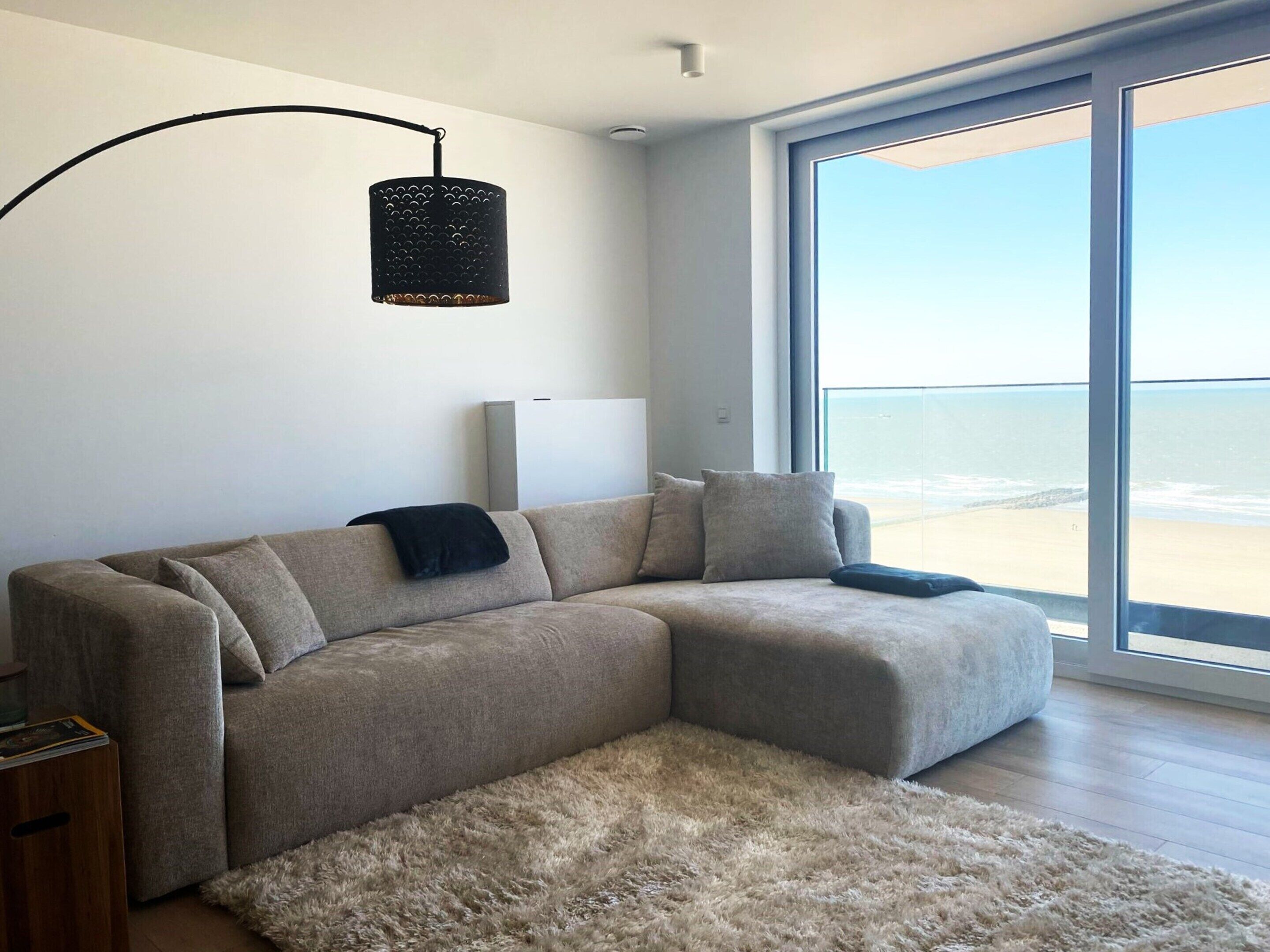 A modern living room with a large sectional sofa, a fluffy rug, a black pendant light, and a large window with a view of the beach.