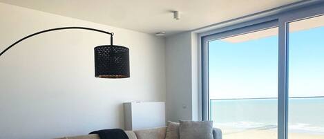 A modern living room with a large sectional sofa, a fluffy rug, a black pendant light, and a large window with a view of the beach.