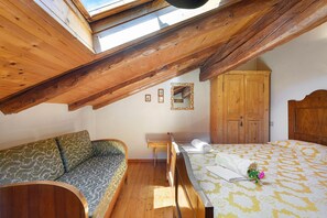 A room with a sloped ceiling, wooden beams, a bed with a patterned bedspread, a sofa with a leaf-patterned cushion, and a wooden wardrobe.