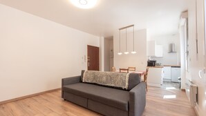 A modern living room with a sofa, a dining area with chairs, a kitchen with appliances, and a wooden floor.
