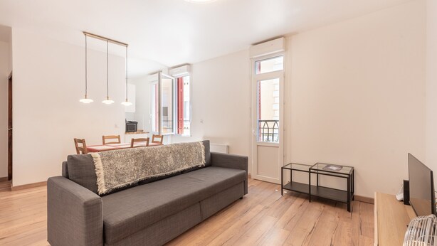 A modern living room with a grey sofa, wooden flooring, a small TV, and a dining area with a table and chairs.