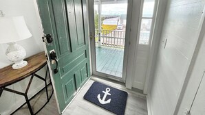A door with a doormat featuring an anchor symbol, a wooden table with a globe and lamp, and a view of a balcony with a railing and a car outside. - Historical Mattocks House - Waterfront, Downtown Swansboro, Boat Slip (Swansboro)